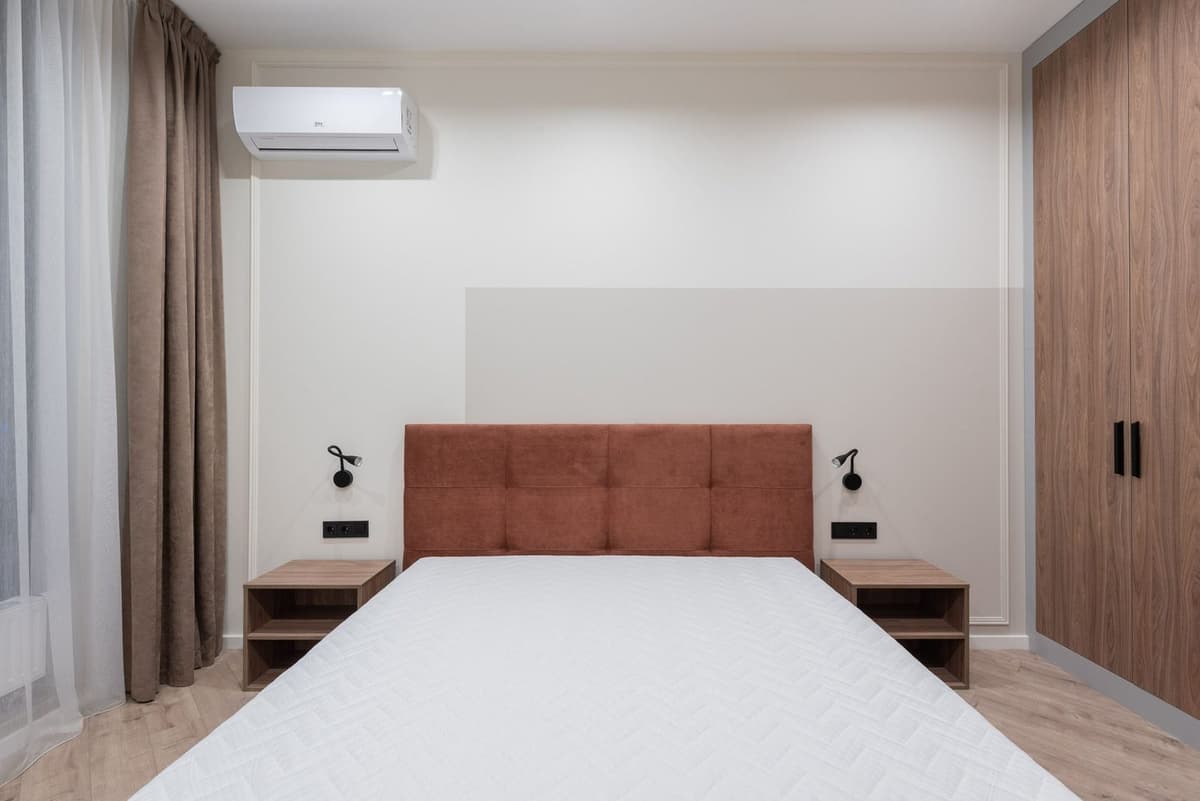 White wall-mounted air conditioner installed above a double bed in a light-toned bedroom