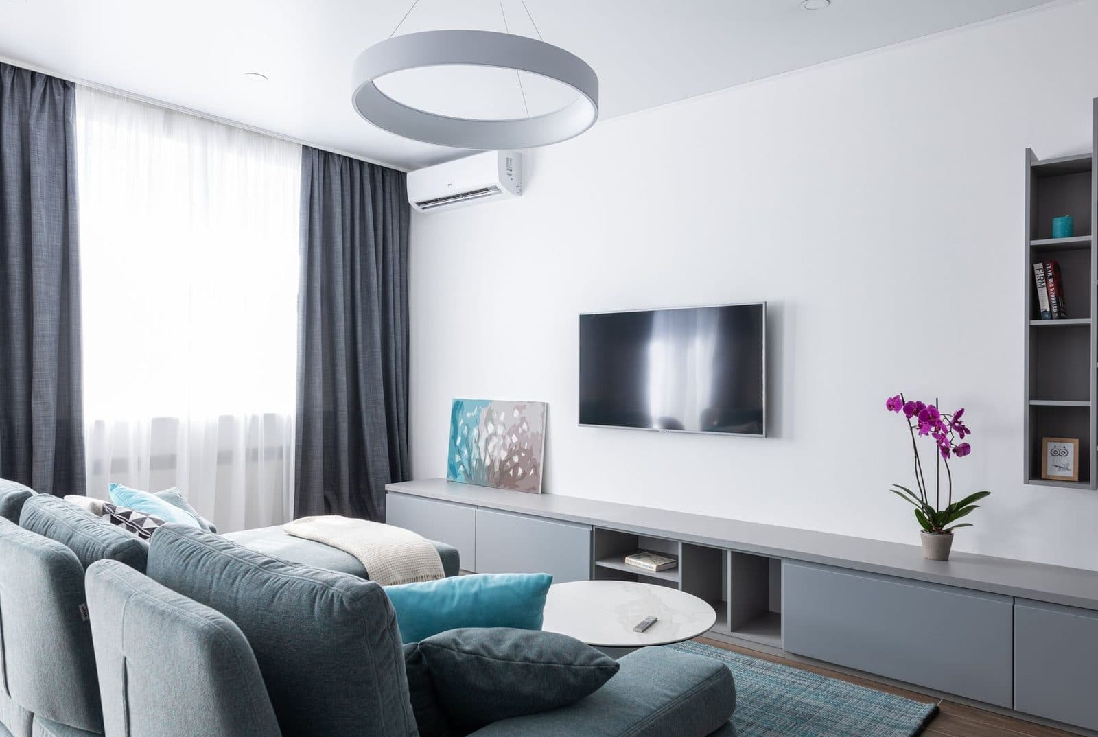 White wall-mounted air conditioner installed in a modern living room with a blue sofa, television and grey curtains