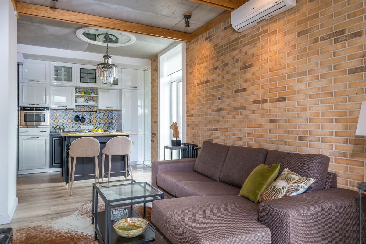 Ar condicionado de parede instalado acima de uma parede de tijolo numa sala de estar luminosa com sofá e cozinha aberta
