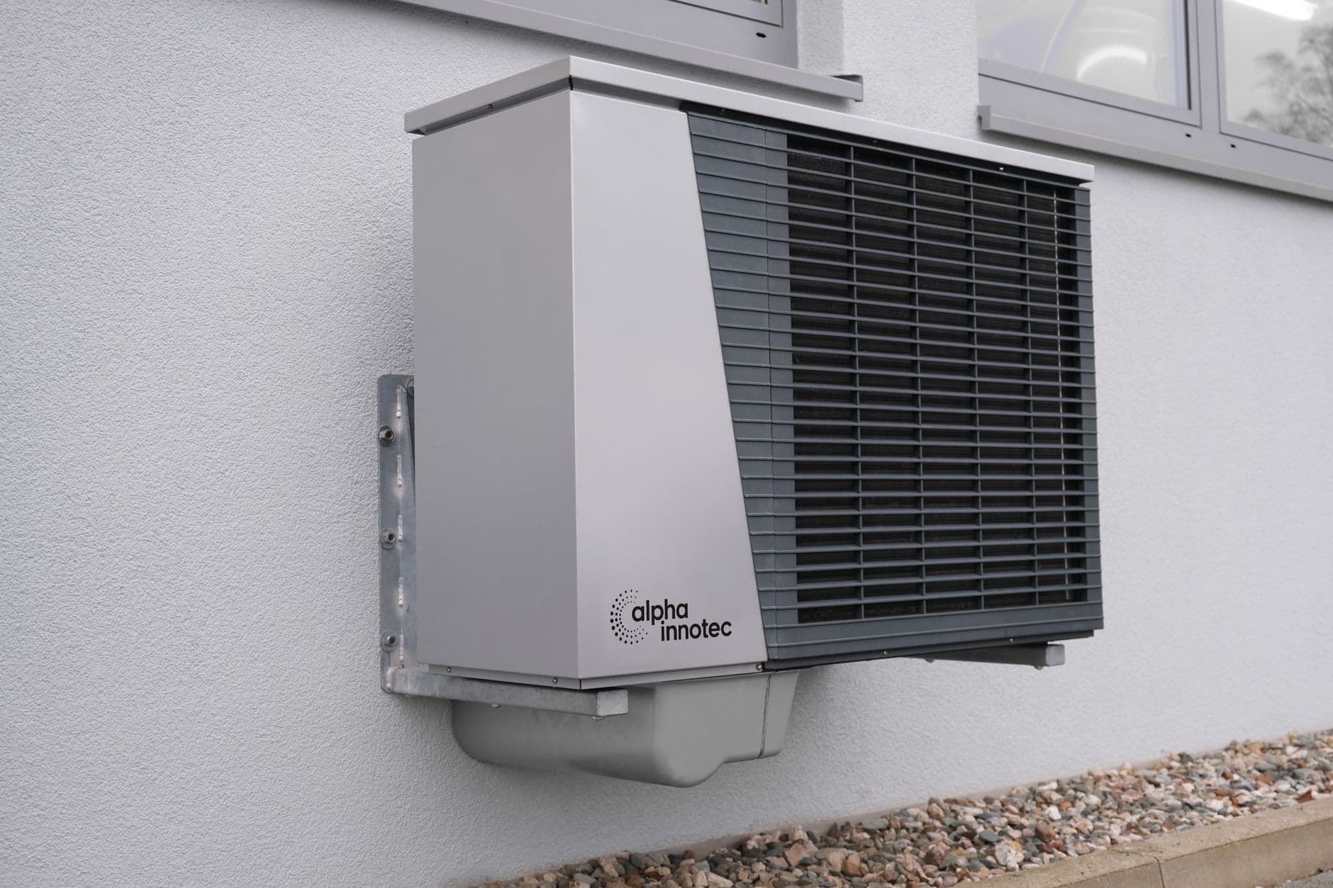 Unidade externa de bomba de calor cinza fixada em uma parede branca com grade de ventilação preta.