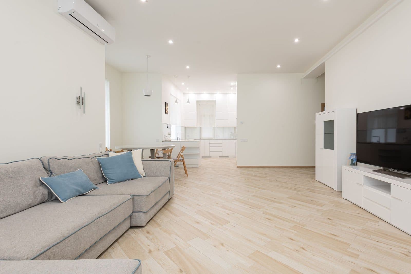 Ar condicionado de parede instalado numa sala de estar luminosa com sofá cinzento, piso de madeira clara e cozinha aberta