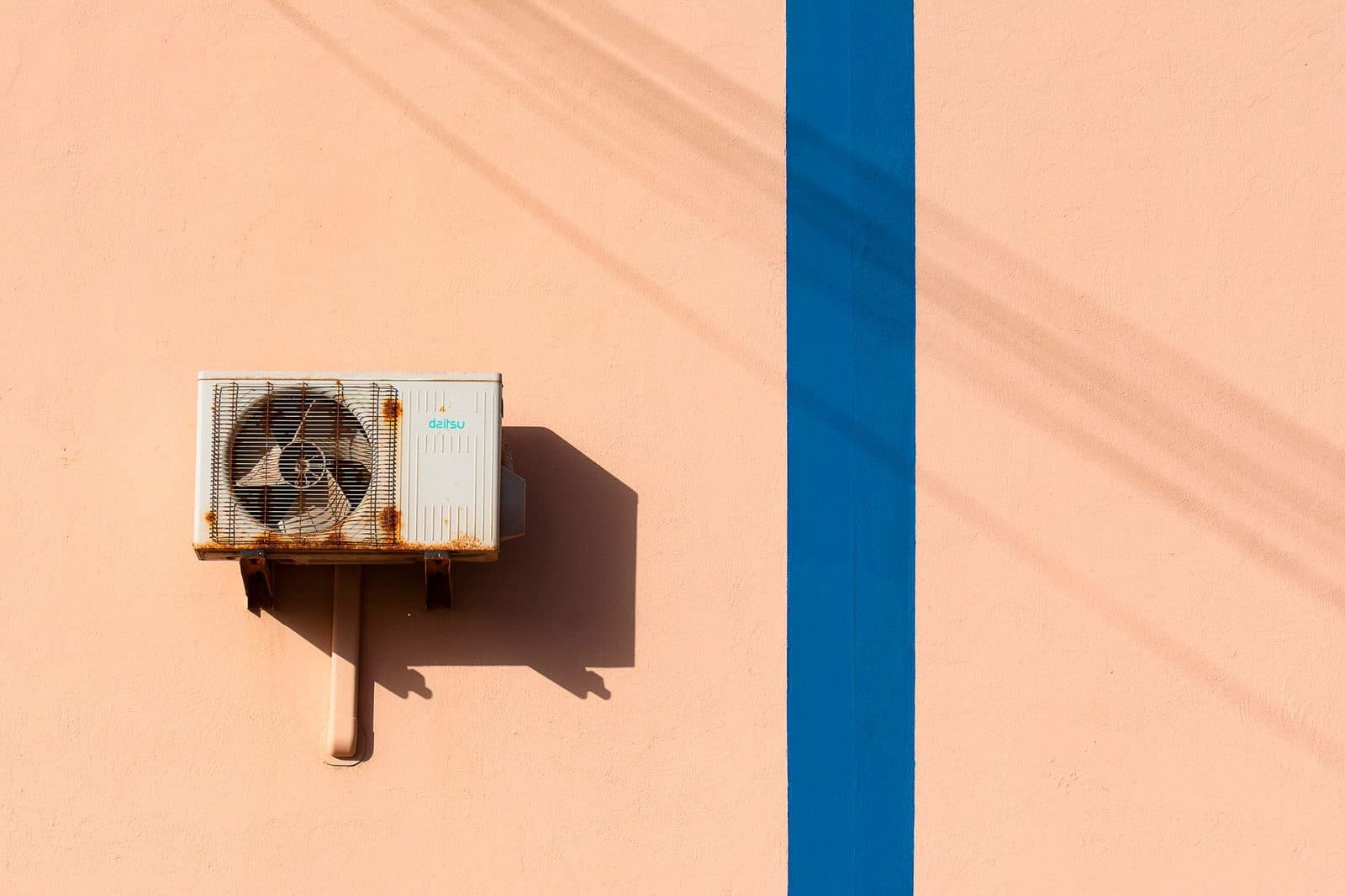 Unité extérieure de climatisation fixée sur un mur rose, à côté d'une bande bleue verticale