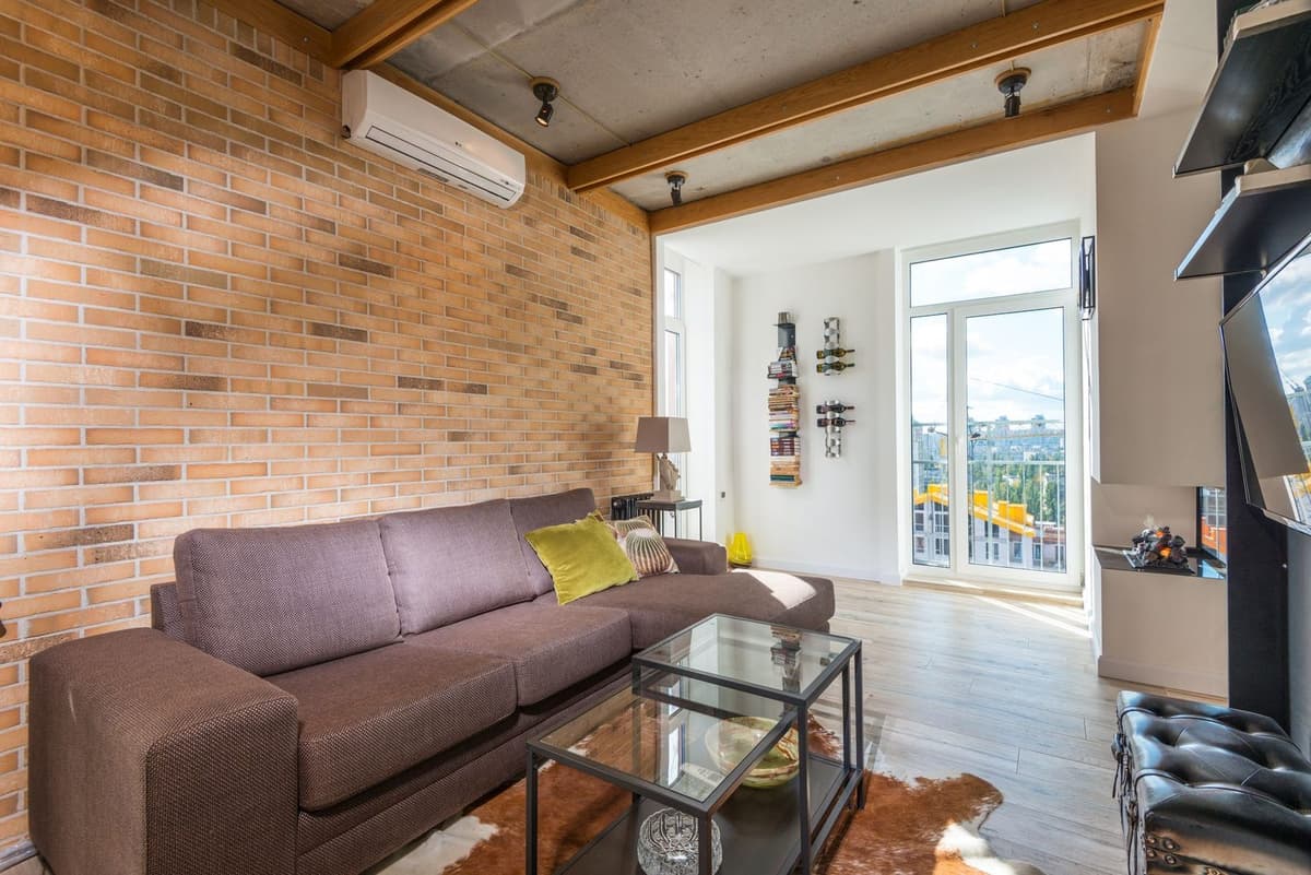 Wandairco gemonteerd op een bakstenen muur in een lichte woonkamer met bank en raampartij
