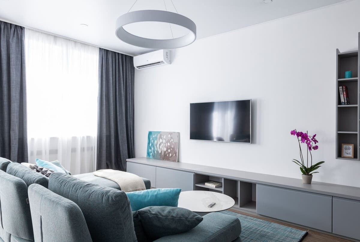 Climatizzatore a parete bianco installato in un soggiorno moderno con divano blu, televisore e tende grigie