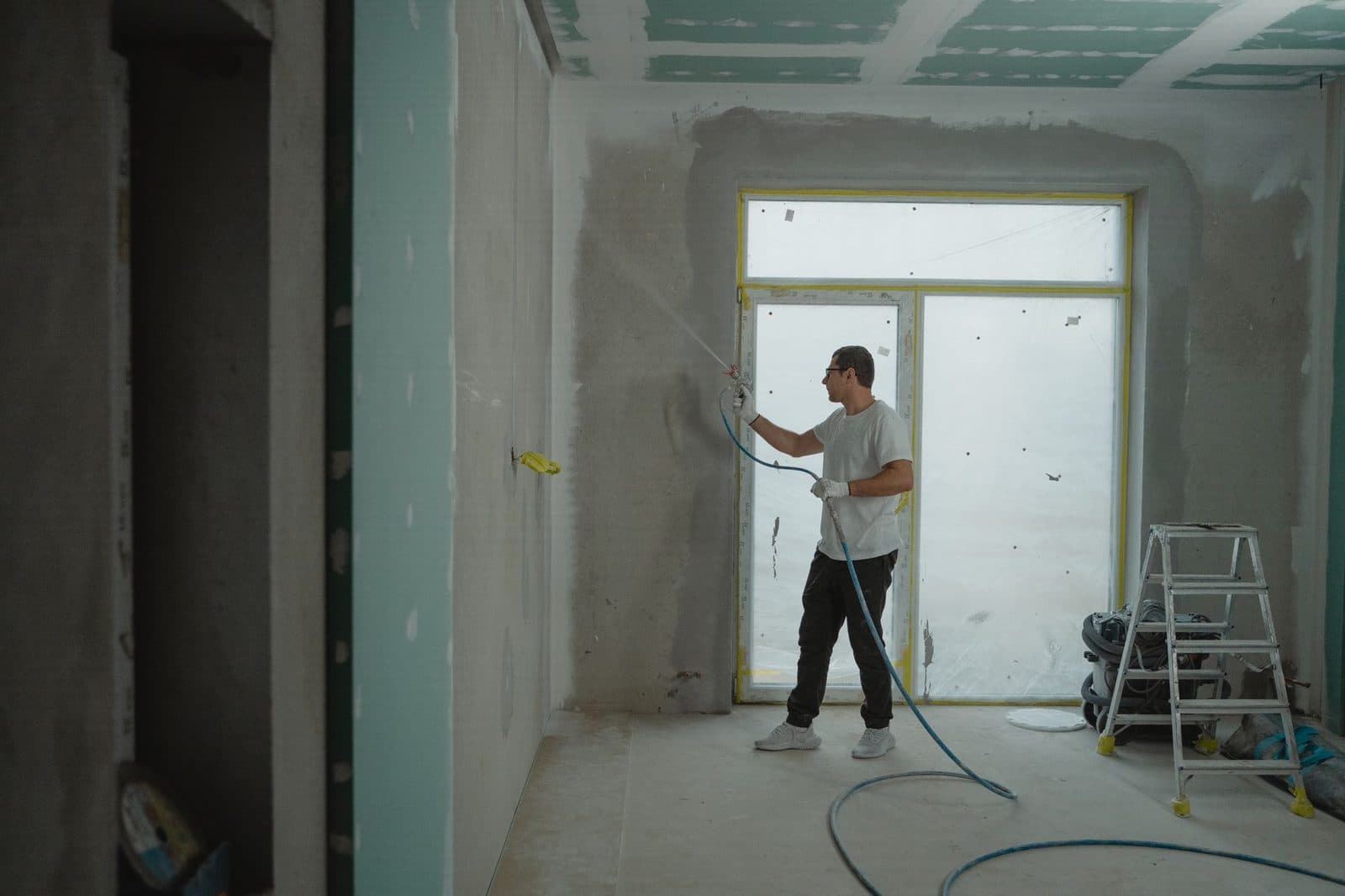 Vakman die pleisterwerk op een muur spuit in een kamer die wordt gerenoveerd, trapje bij het raam