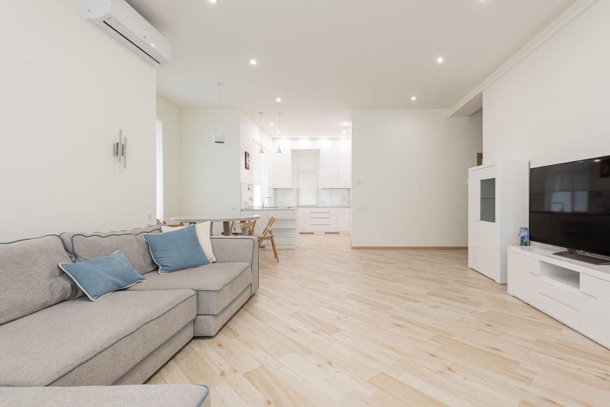 Ar condicionado de parede instalado numa sala de estar luminosa com sofá cinzento, piso de madeira clara e cozinha aberta