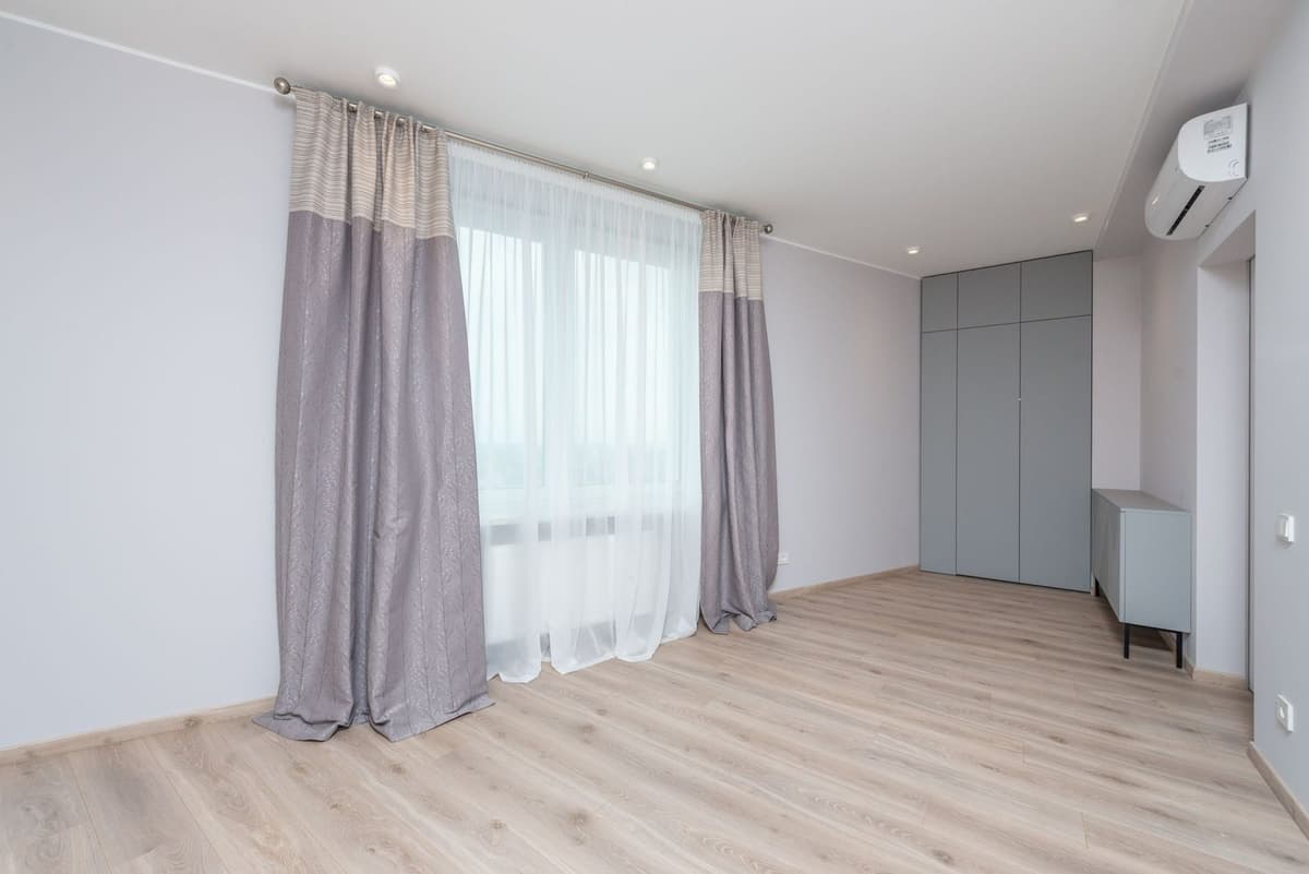 Aire acondicionado de pared instalado en alto en una habitación vacía con paredes claras, suelo de parqué y cortinas grises