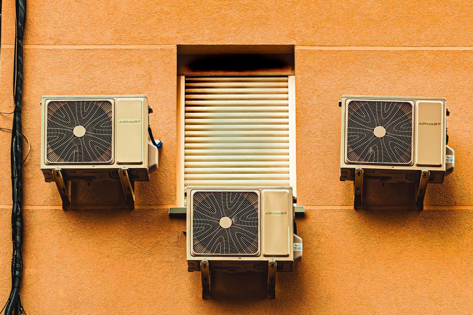 Trois unités extérieures de climatisation fixées sur un mur ocre d'immeuble, près d'un volet roulant