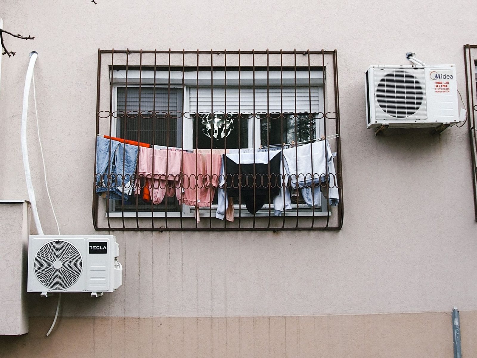 Dos unidades exteriores de aire acondicionado fijadas en la fachada de un edificio, junto a una ventana con reja y ropa tendida.