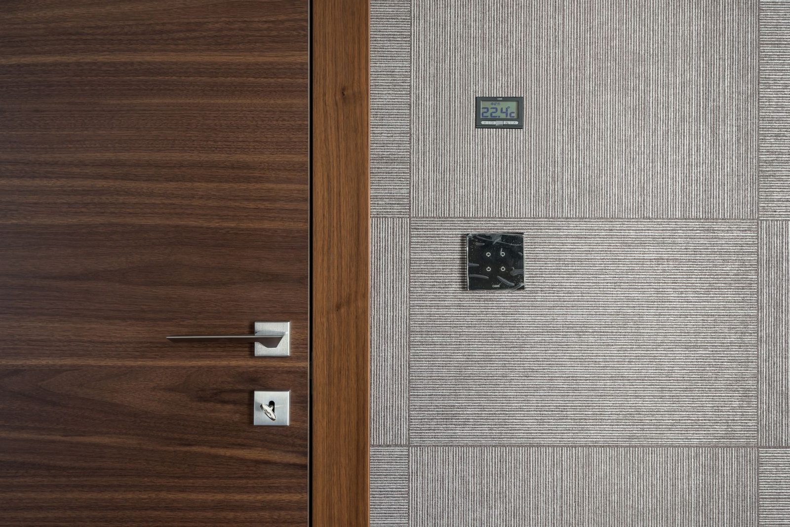 Wandthermostat mit Anzeige 22,4 °C und schwarzer Schalter auf grau gefliester Wand, neben einer Holztür