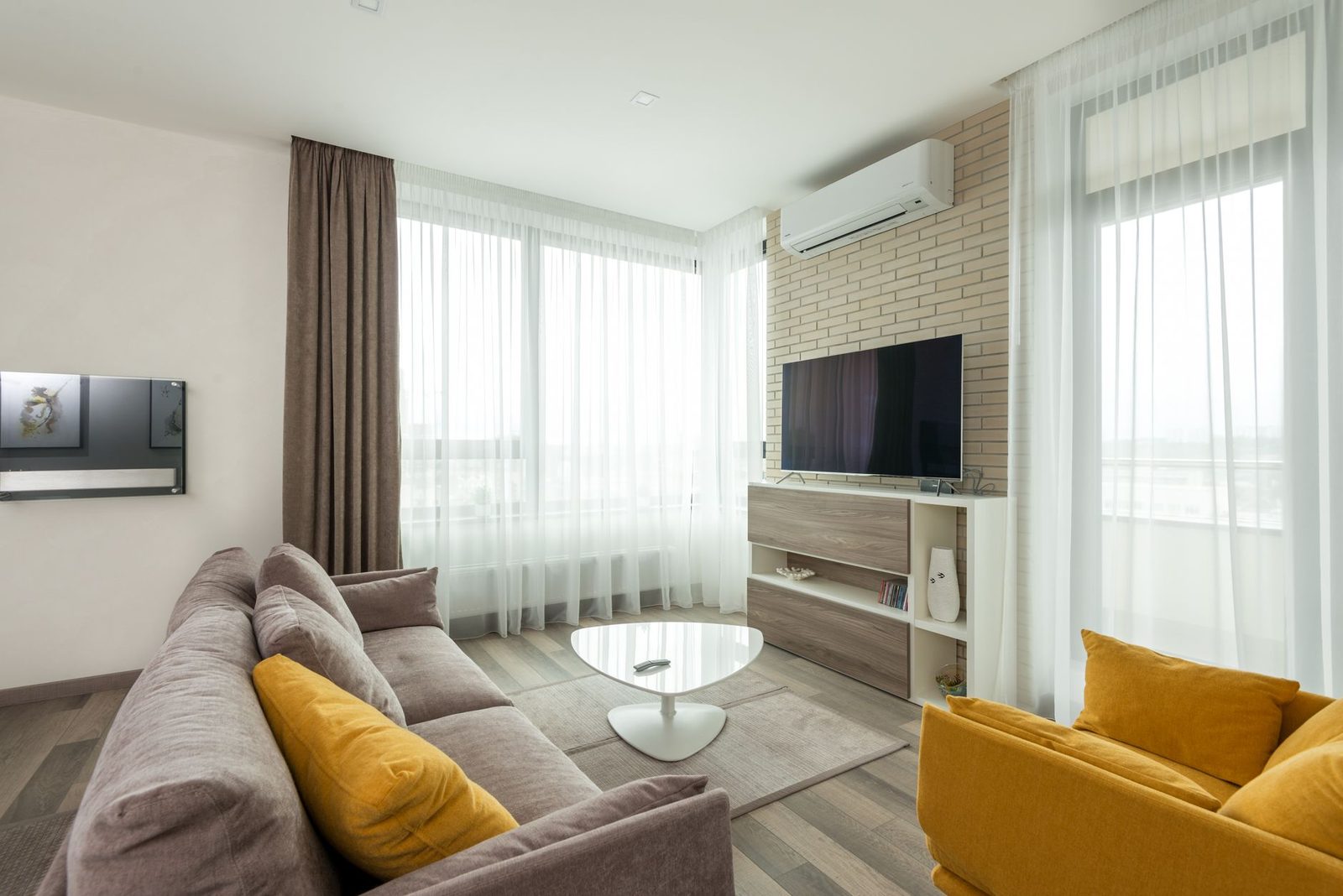 White wall-mounted air conditioner installed above a television in a bright living room with grey and yellow sofas