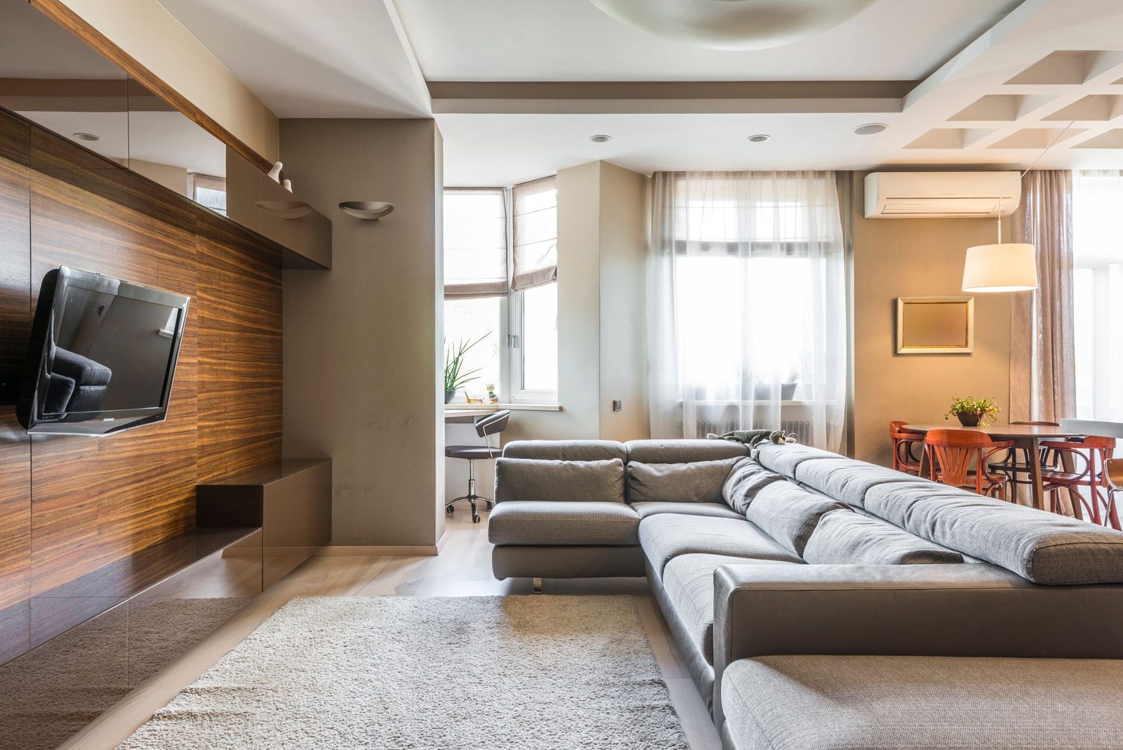 White wall-mounted air conditioner installed high up in a modern living room with a grey corner sofa and a television