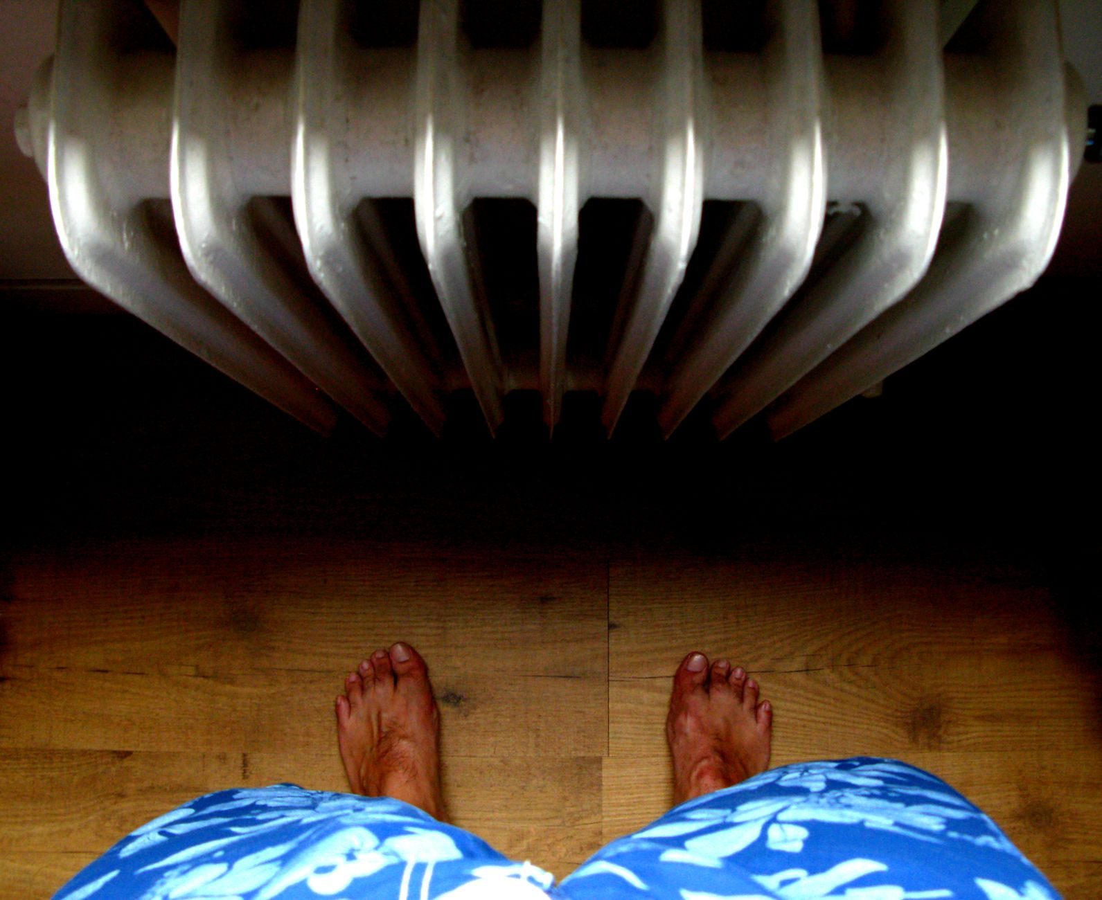 Blote voeten op een parketvloer voor een witte gietijzeren radiator, van bovenaf gezien