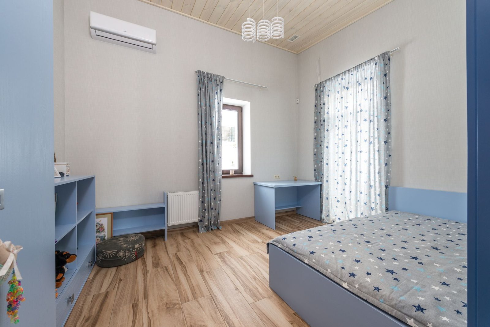 Unidad mural de aire acondicionado en una habitación infantil con muebles azules, suelo de parqué claro y cortinas de estrellas
